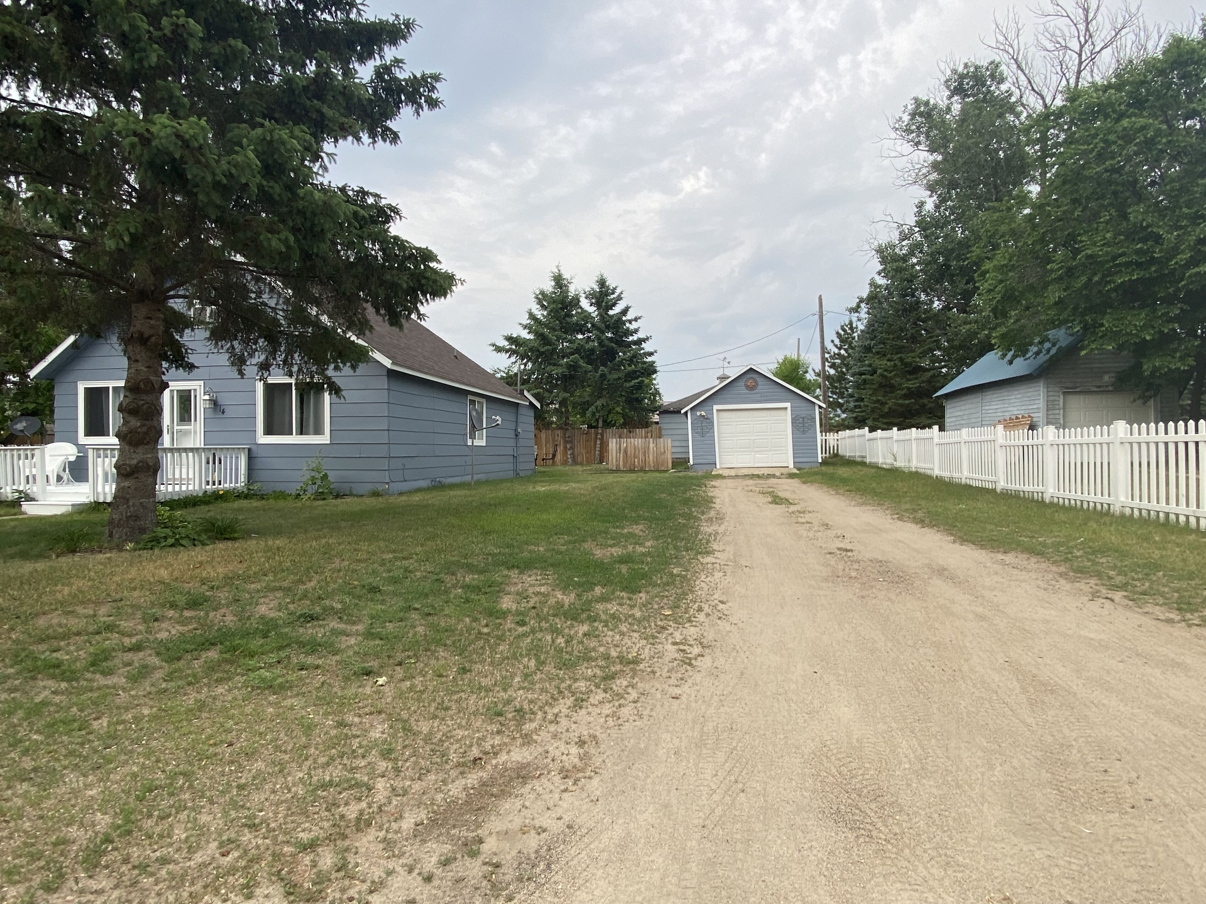 A rural driveway leading to a house with a garage, surrounded by a white picket fence and trees.