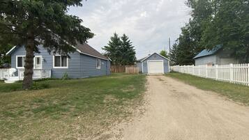 A rural driveway leading to a house with a garage, surrounded by a white picket fence and trees.
