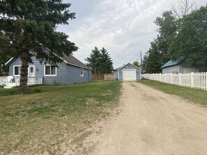 Un chemin de terre menant à une maison avec un garage, entourée d’une clôture en bois peinte en blanc et d’arbres.