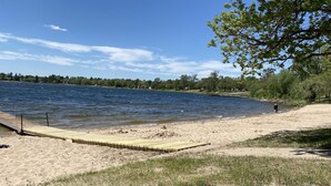 Ein sandiger Strand mit einem hölzernen Steg, der in einen See führt, umgeben von Bäumen und einem klaren Himmel.