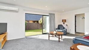 A modern living room with a flat-screen TV, a blue sofa, a wooden coffee table, and a sliding glass door leading to a backyard.