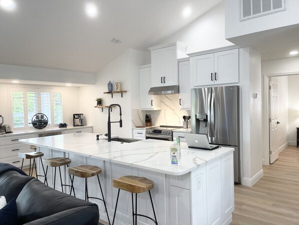 A modern kitchen with white cabinetry, a stainless steel refrigerator, a built-in oven, and a large island with bar stools.