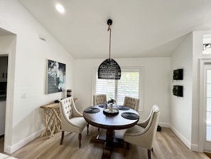 A dining area with a round wooden table, four upholstered chairs, a hanging black chandelier, and a painting on the wall.