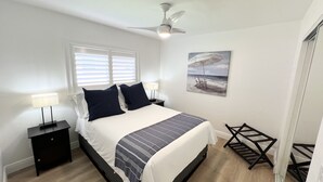 Ein Schlafzimmer mit einem Bett, einem Deckenventilator, einem Nachttisch mit Lampe, einem Bild einer Strandansicht und einem Holzboden.