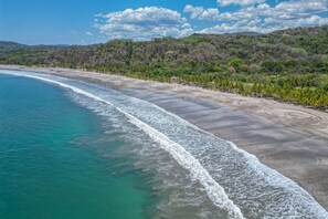 Ein Strand mit klarem türkisblauem Wasser, einem sandigen Ufer und einem bewaldeten Hügel im Hintergrund.
