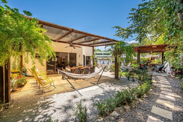 Ein überdachter Patio-Bereich mit Esstisch und Stühlen, einer Hängematte und einer Pergola mit Bepflanzung.