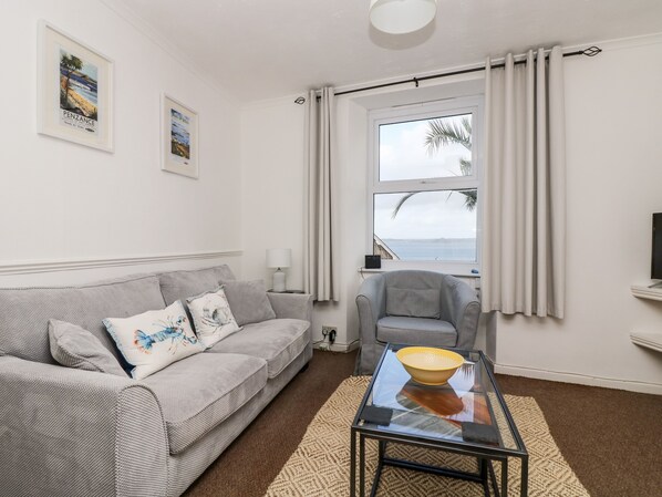 A living room with a grey sofa, a glass coffee table, a small armchair, and a view of the sea through a window.