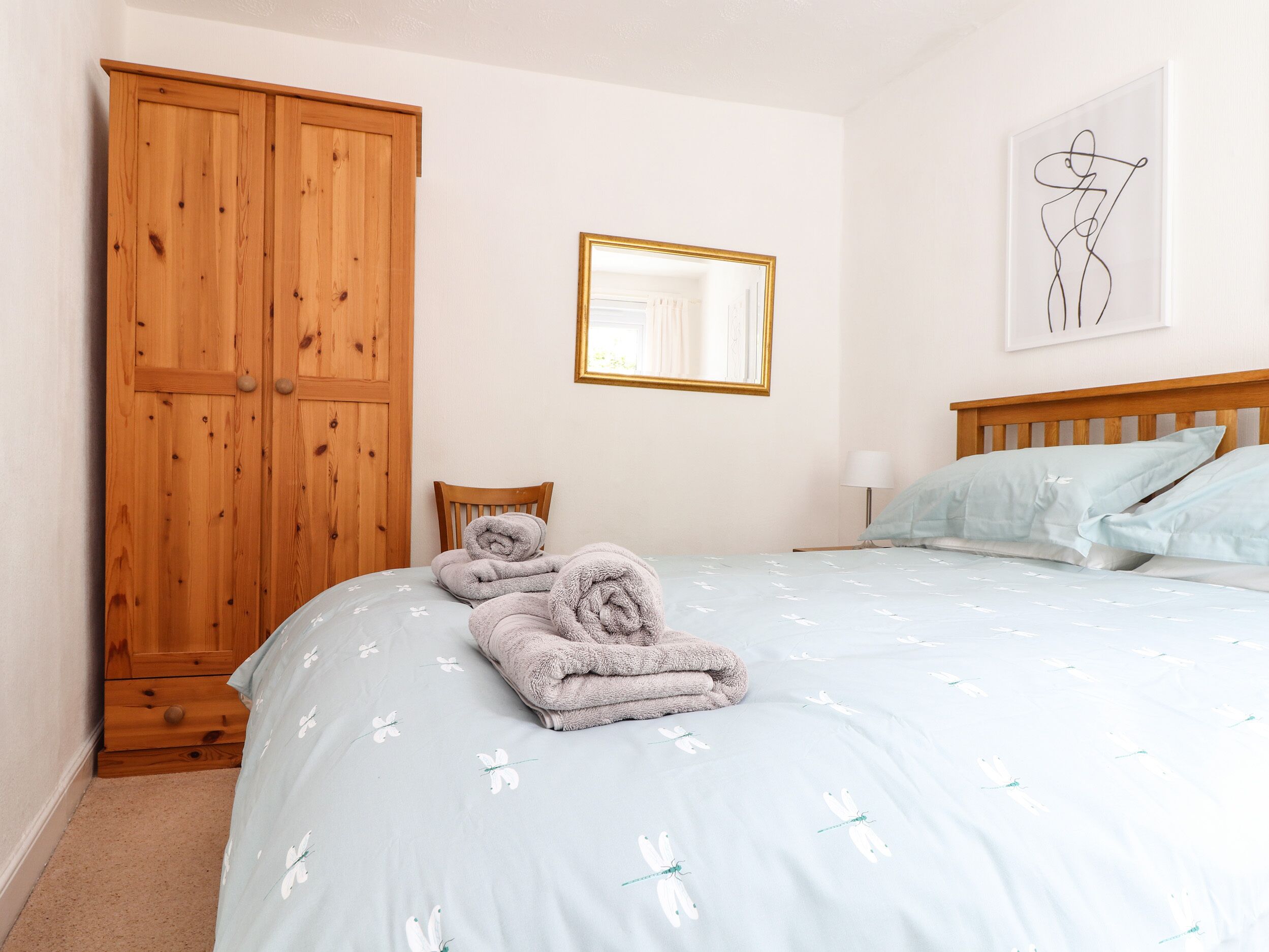 Une chambre à coucher comprenant un lit, une armoire en bois, une chaise, une table de chevet avec une lampe et une œuvre d’art encadrée accrochée au mur.