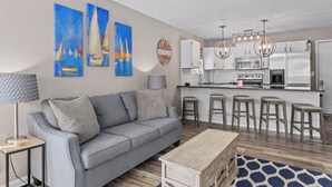 Una sala de estar moderna con un sofá gris, una mesa de centro y una cocina con armarios blancos y electrodomésticos de acero inoxidable.