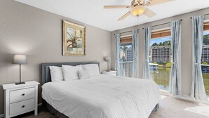 Un dormitorio con cama, mesitas de noche, ventilador de techo, un cuadro y vista a un edificio y al agua.