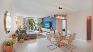 Un apartamento moderno con comedor, sala de estar con televisor grande y balcón con vista a palmeras.
