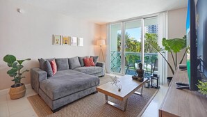 Un salón moderno con un sofá gris, una mesa de centro y un televisor sobre un mueble de madera. Hay un balcón con vistas a palmeras y edificios.
