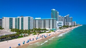 Un hotel frente a la playa con varios edificios de gran altura, una playa de arena con sombrillas y un cielo azul despejado.