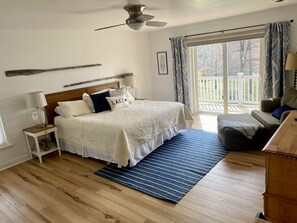 Ein Schlafzimmer mit einem Bett, einem Nachttisch, einer Lampe, einem Deckenventilator, einem Fenster mit Vorhängen und Blick auf eine Terrasse.