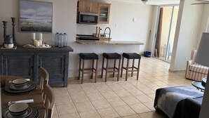 Una cocina moderna con armarios de madera, microondas y una barra con taburetes. Hay una mesa de comedor dispuesta con platos y un cuadro encima.