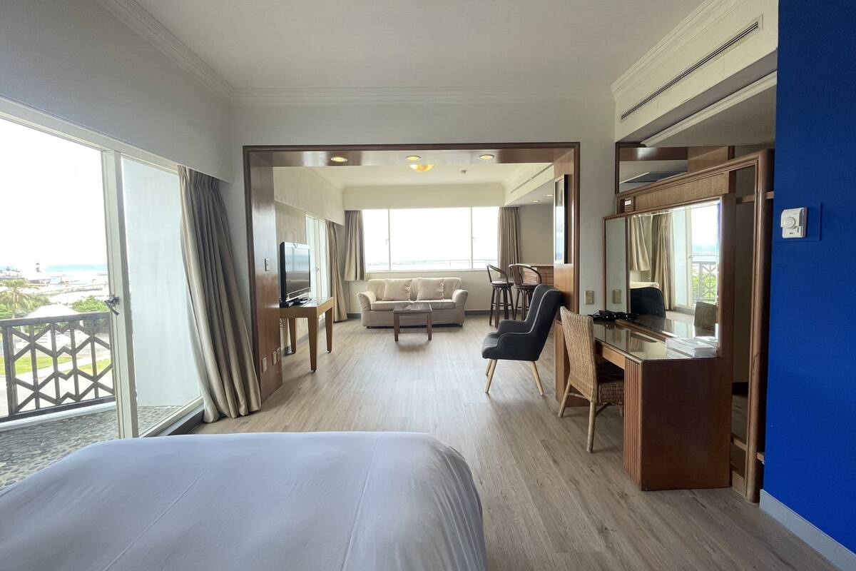 Een hotelkamer met een groot bed, een bureau met stoel, een bank en een televisie. Er is een balkon met uitzicht op de stad.
