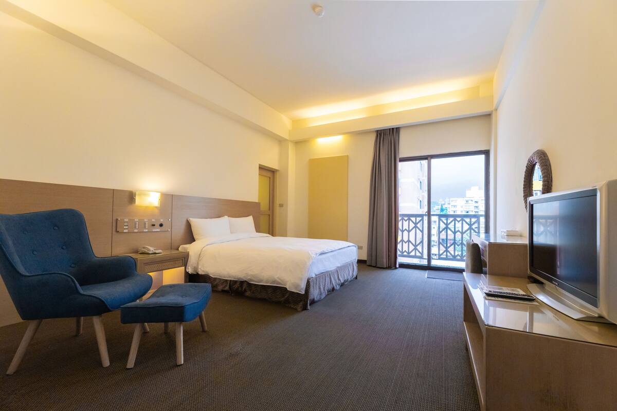 Een hotelkamer met een bed, een blauwe fauteuil, een televisie en uitzicht op de stad.