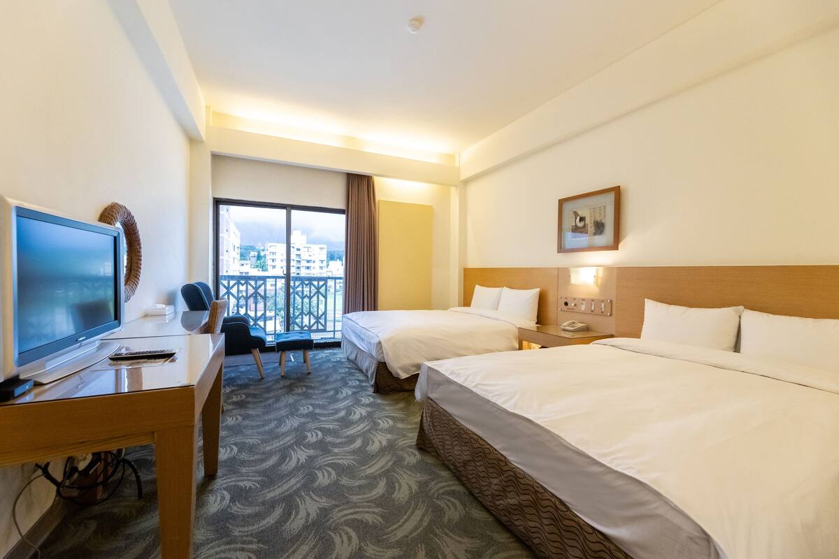 Een hotelkamer met twee bedden, een bureau met een televisie, een stoel en uitzicht op de stad door een groot raam.