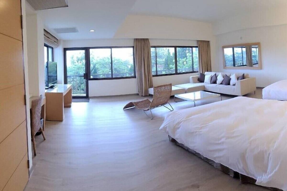 Ruime hotelkamer met een groot bed, een bank, een eettafel en een stoel. Grote ramen bieden uitzicht op het omliggende landschap.