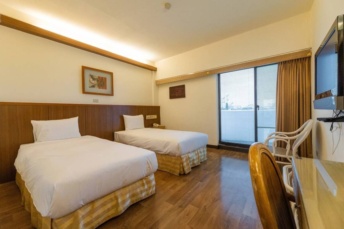 Een hotelkamer met twee bedden, een bureau, een stoel, een televisie en een groot raam met gordijnen.