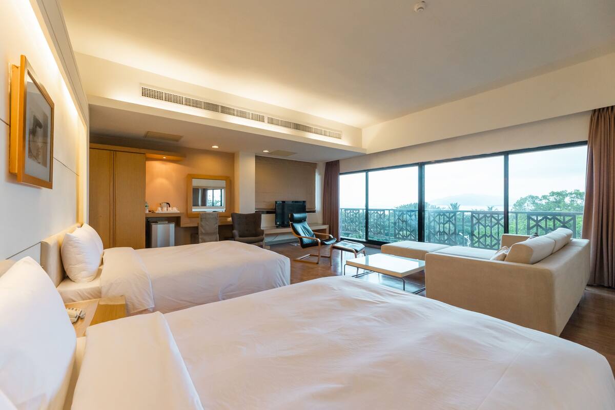 Hotelkamer met twee bedden, een slaapbank, een bureau met stoel en een groot raam met uitzicht op het landschap.