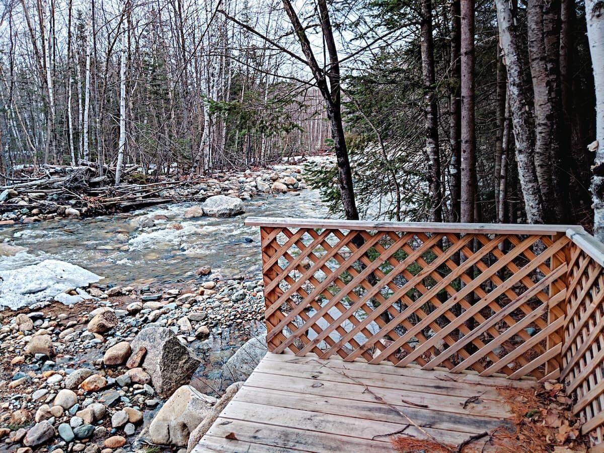 A wooden deck with a lattice railing overlooking a rocky riverbed and forested area.