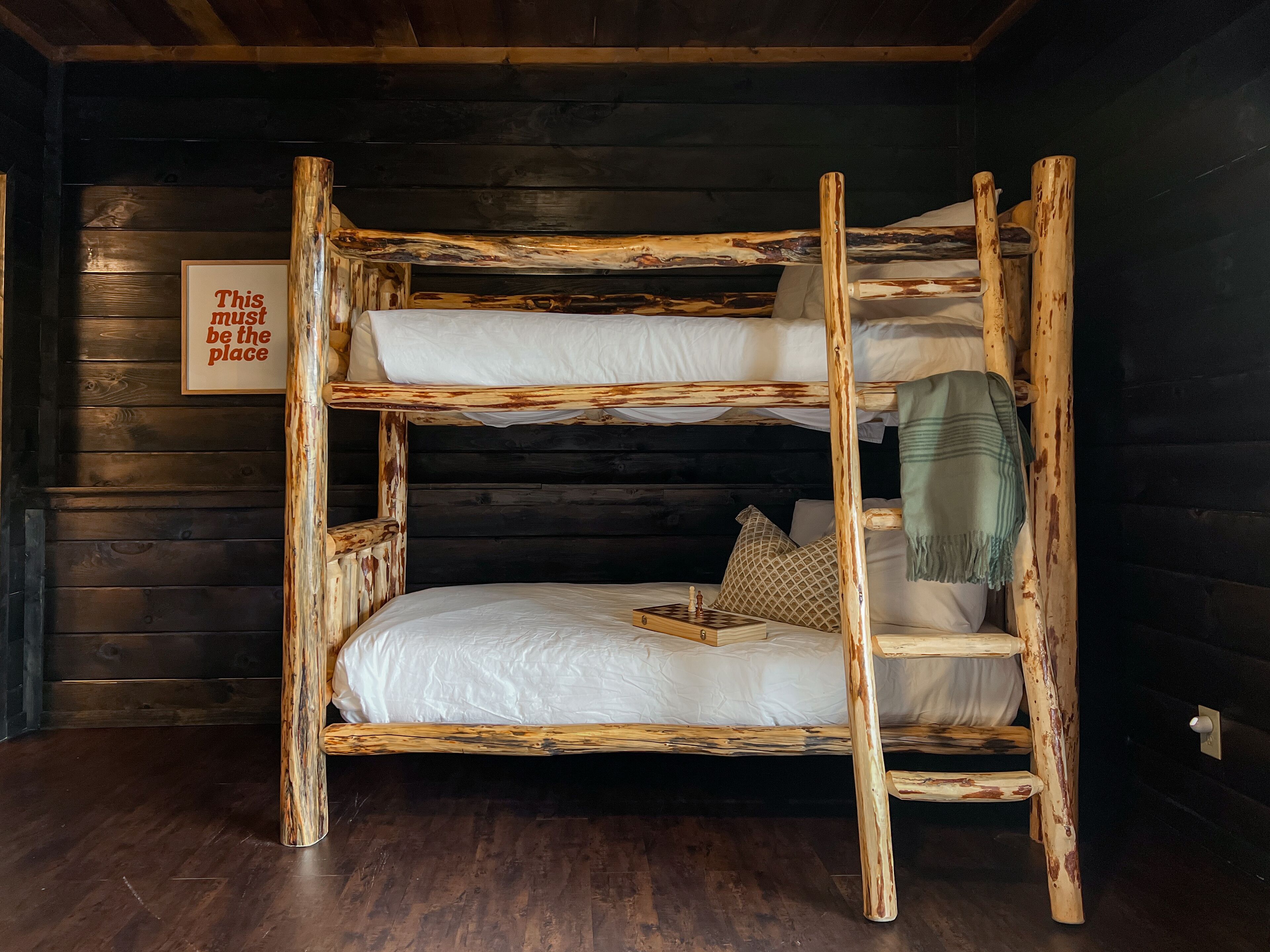 Un lit superposé rustique avec une structure en bois, une literie blanche et un jeu d’échecs posé sur le lit du bas.