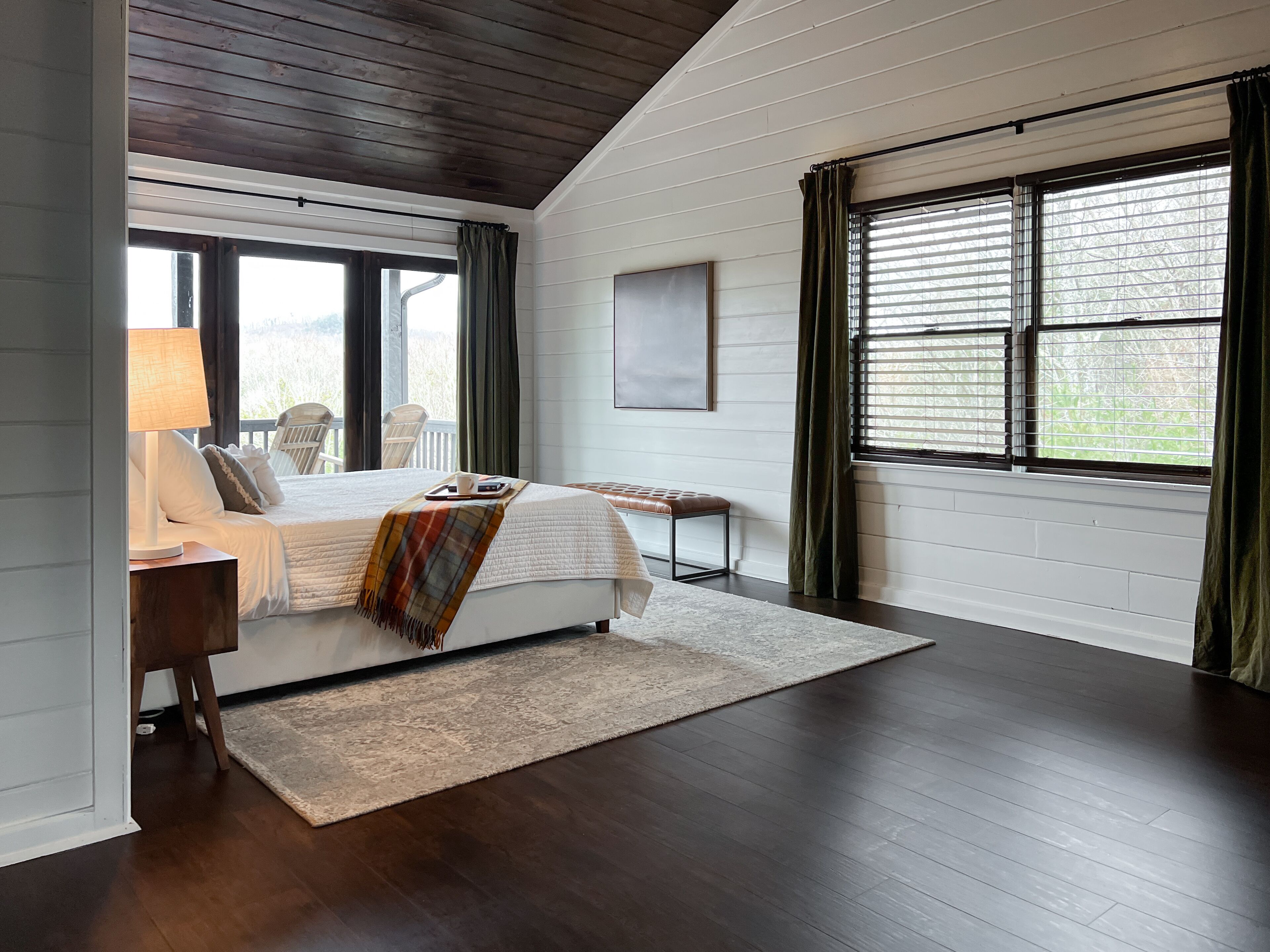 A bedroom with a bed, bedside table, lamp, chair, bench, and large windows with curtains.