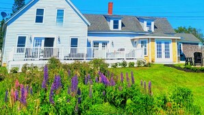 A two-story house with a white exterior, a porch with a white railing, and a small garden with purple flowers in front. - Spectacular Corea Harbor side Home minutes to Acadia National Park Schoodic Gate (Gouldsboro)