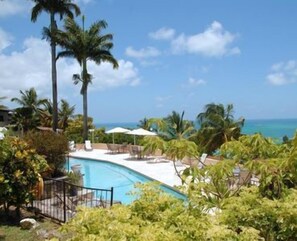 A pool area with lounge chairs and umbrellas, surrounded by palm trees and a view of the ocean. - Swimming Pool- Balcony/Veranda- Sea & Sunset Views (St Johns)