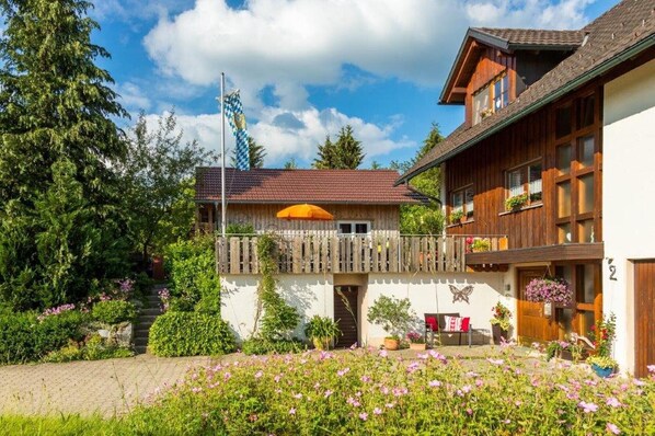 A two-story wooden chalet with a balcony, surrounded by lush greenery and flowering plants.