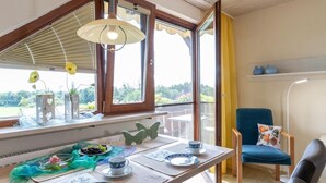 Un coin repas avec une table dressée pour deux personnes, une chaise avec un coussin et une vue sur la verdure environnante.