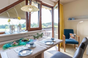 A dining area with a table set for two, a chair with a cushion, and a view of greenery outside.