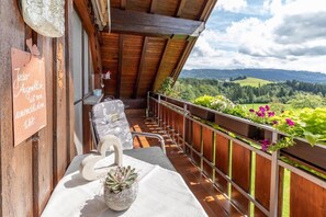 A balcony with a wooden railing, a chair, a swan-shaped cushion, a potted succulent, and a view of a green landscape.