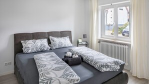 A bed with a grey mattress and patterned pillows, a bedside table with a lamp, and a window with a view of a car and greenery. - Pet-Friendly Apartment Holly with Wi-Fi & Terrace (Ebringen)