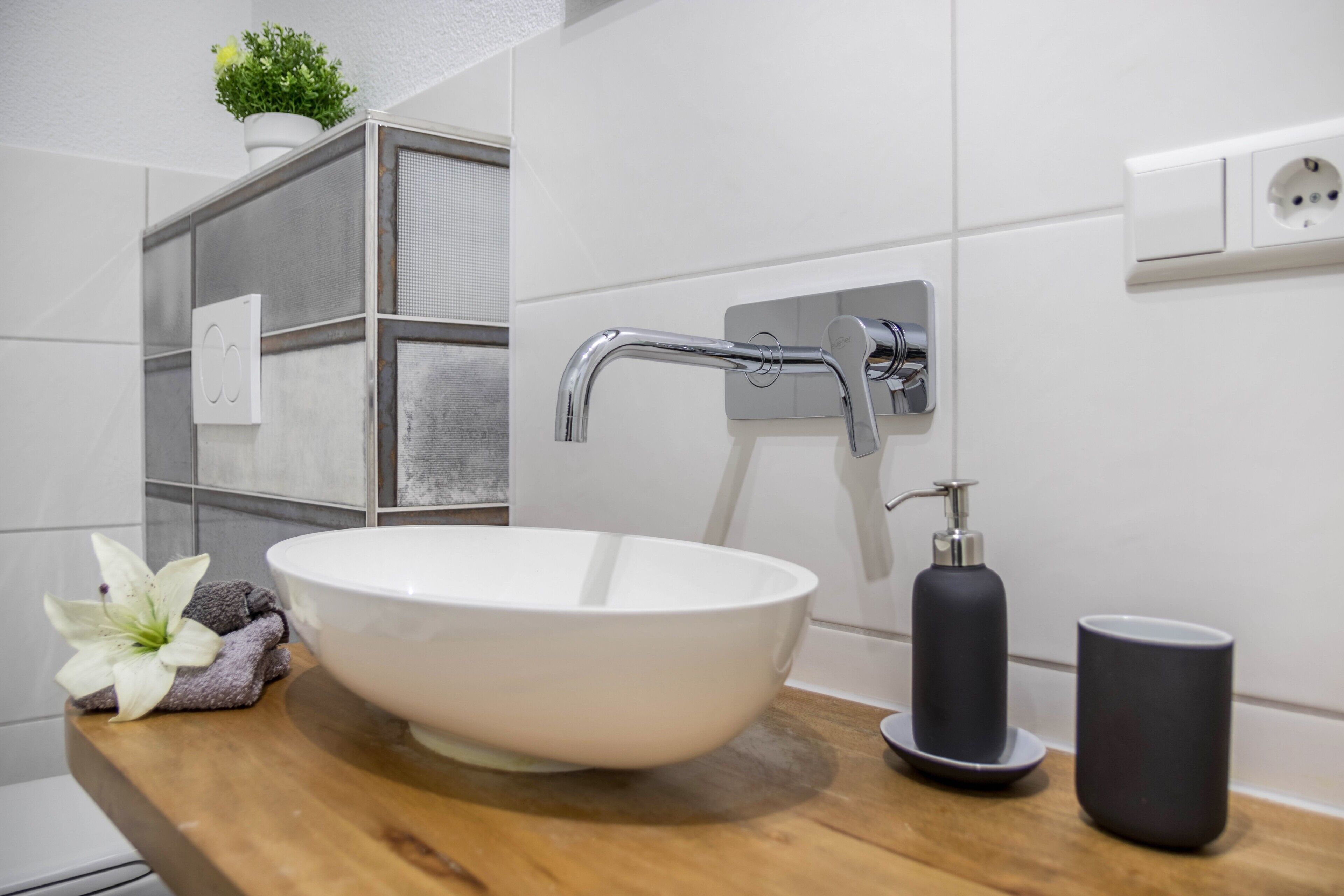 Une salle de bain avec un lavabo sur colonne blanc, un plan de toilette en bois, une serviette, une fleur, un distributeur de savon et un gobelet.