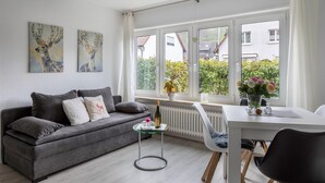 A modern living room with a grey sofa, a glass coffee table, a white dining table, and a window with a view of greenery and buildings. - Pet-Friendly Apartment Holly with Wi-Fi & Terrace (Ebringen)
