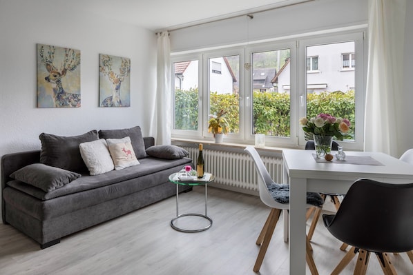 A modern living room with a grey sofa, a glass coffee table, a white dining table, and a window with a view of greenery and buildings.