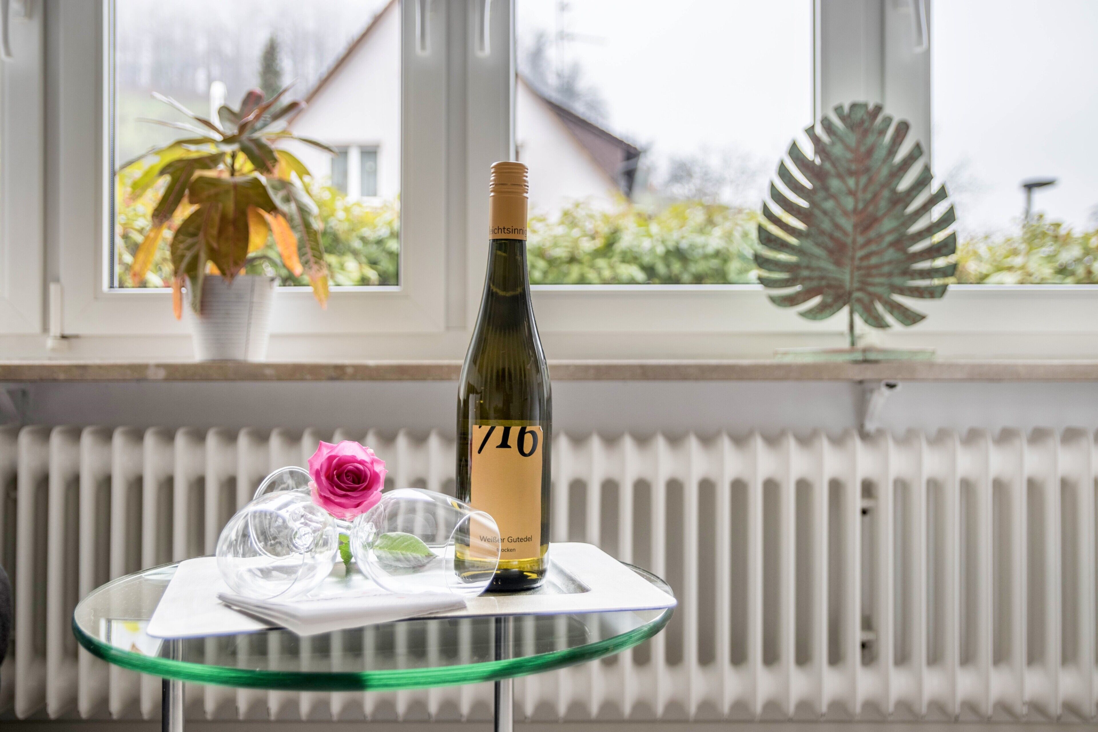Una botella de vino sobre una mesa de vidrio, con una planta en maceta y una hoja decorativa al fondo.