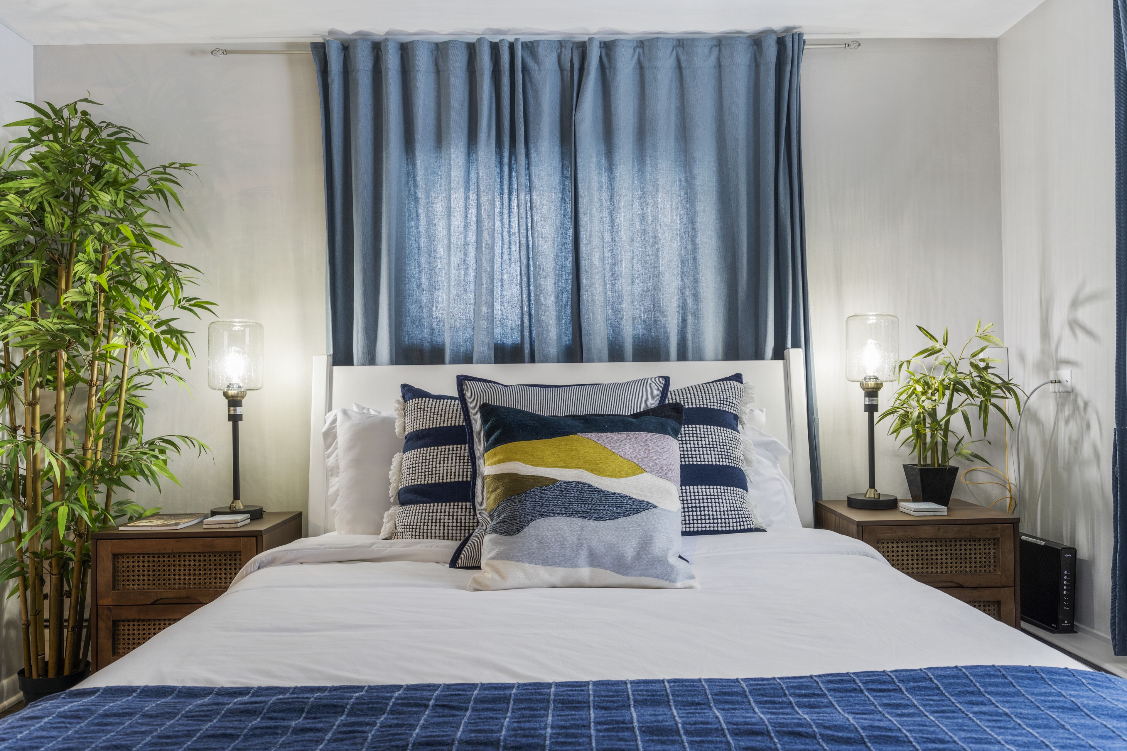 A neatly made bed with a blue blanket, two bedside tables with lamps, and a large window with blue curtains.