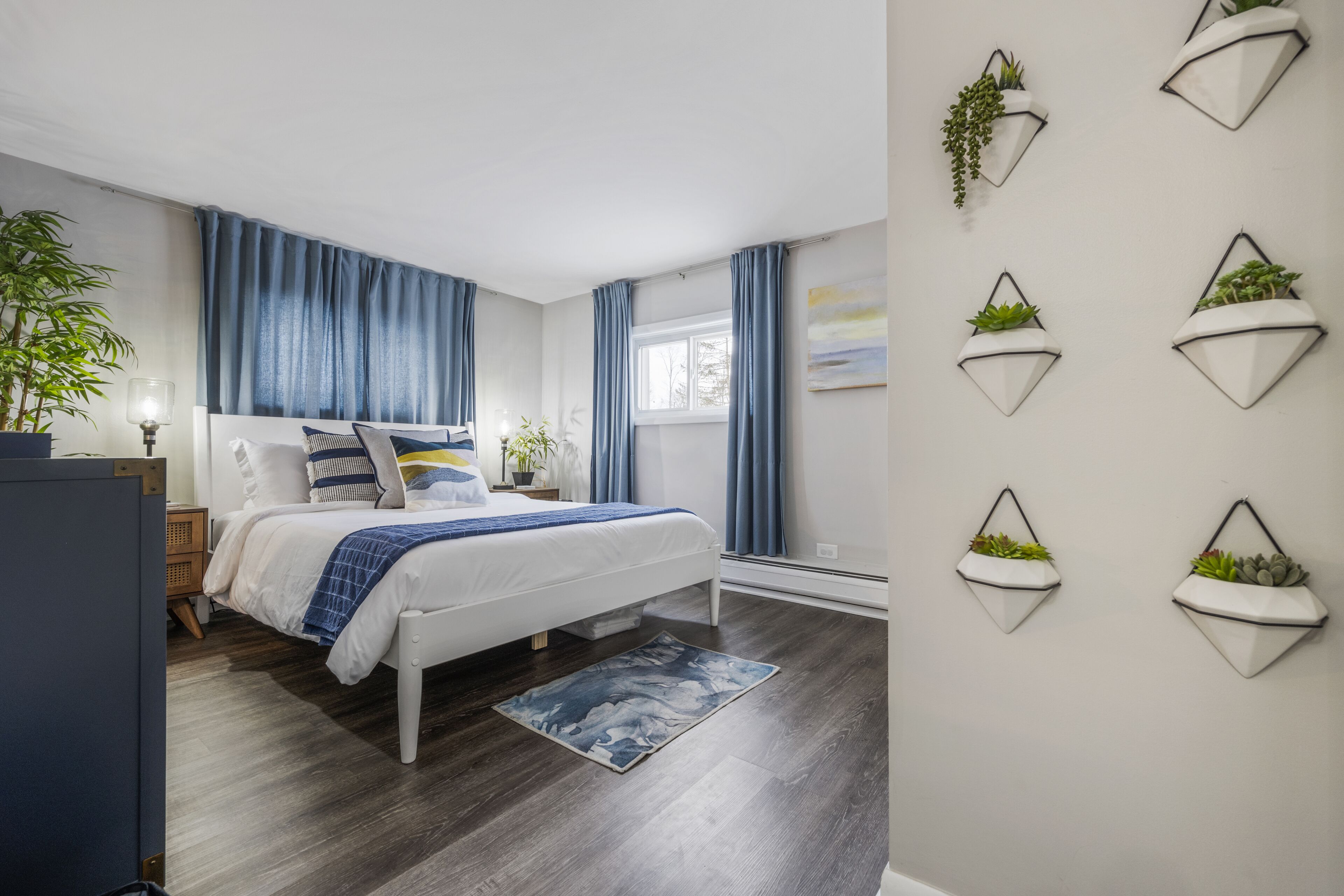Ein modernes Schlafzimmer mit Bett, Nachttischen, einer Lampe, einem Topf mit Pflanze und einem Fenster mit Vorhängen.