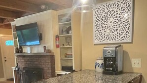 Une cuisine dotée d’un comptoir en granit, de tabourets de bar en bois, d’une machine à café, d’un téléviseur fixé au-dessus du comptoir et d’un élément décoratif mural.