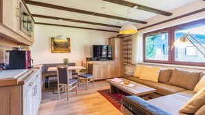 A cozy living room with a sofa, a dining table with chairs, a kitchen area with a TV, and a large window with a view of a house.