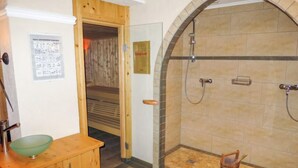 Un baño con un mueble de madera, un cuenco verde, una ducha con pared de azulejos de piedra y una puerta de madera que da a una sala de sauna.