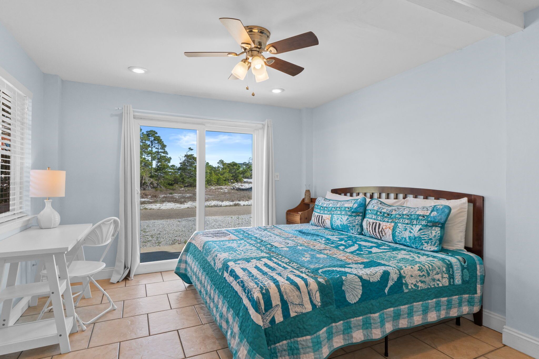 Une chambre à coucher comprenant un lit, un bureau, une chaise, un ventilateur de plafond et une vue sur l’extérieur, visible à travers une porte vitrée.