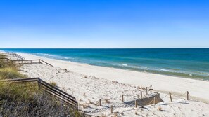A beach with a wooden walkway leading to the water, a sandy area with some vegetation, and a clear blue sky.
