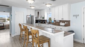 Une cuisine moderne avec des meubles blancs, un grand îlot central doté de tabourets, des appareils en acier inoxydable et un éclairage suspendu.
