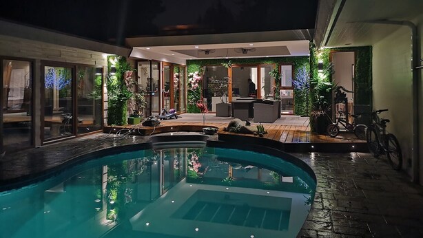 Un espace piscine intérieure doté de murs en verre, d’une terrasse en bois et d’un coin salon avec une table et des chaises.