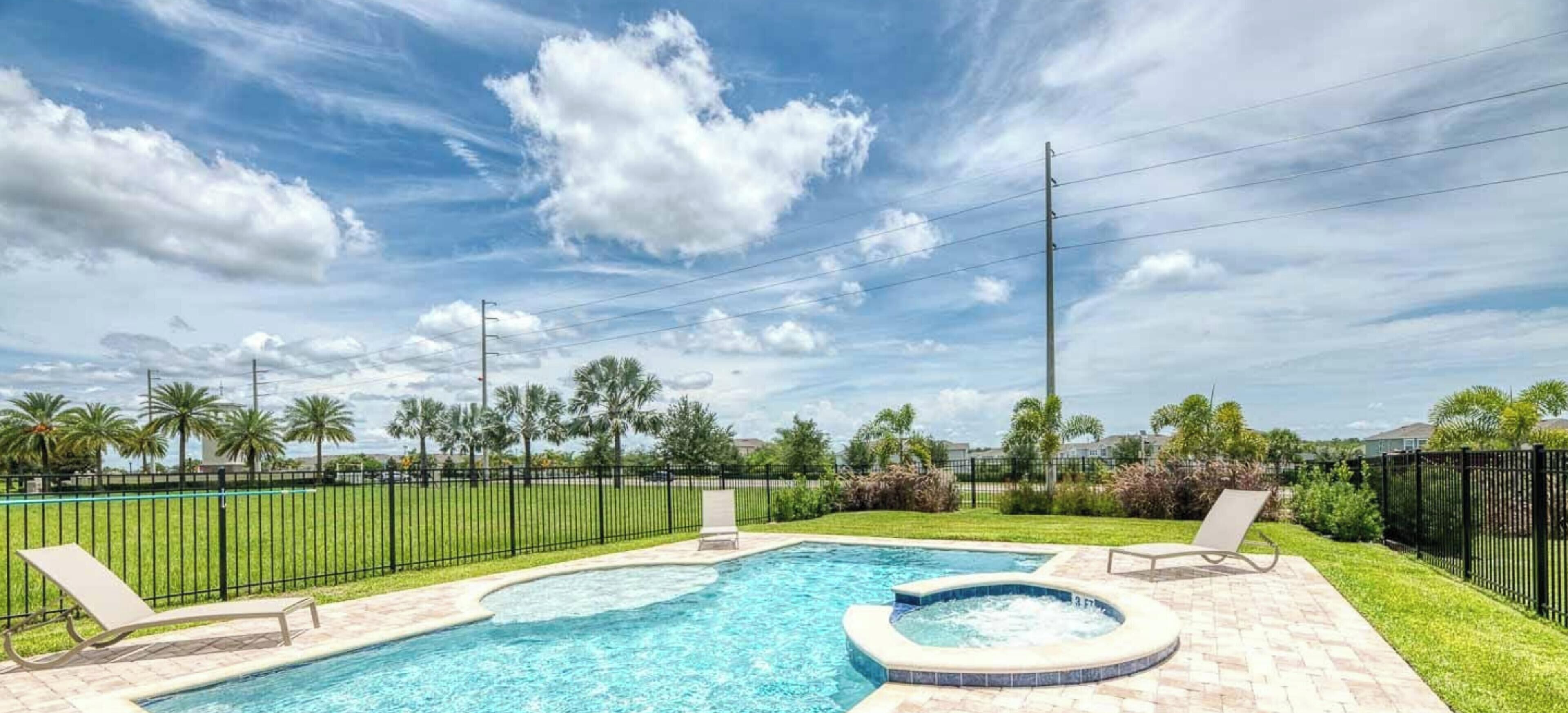 A swimming pool with two lounge chairs and a hot tub, surrounded by a fence and palm trees.