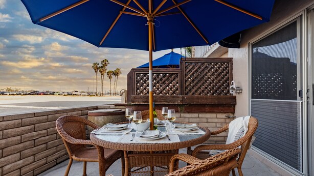 A round table with wicker chairs, a blue umbrella, and a grill set up on a patio overlooking the beach. - Steps to the Sand + Patio Lounging | Sunset Water Views | Bay View 3 (San Diego)